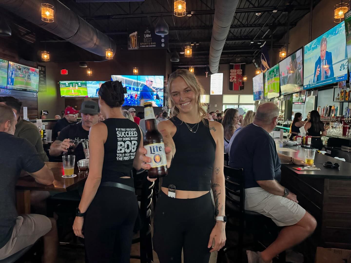 Smiling server holding a Miller Lite at Ed's Tavern sports bar in Bradenton with HD TVs and a full crowd in the background